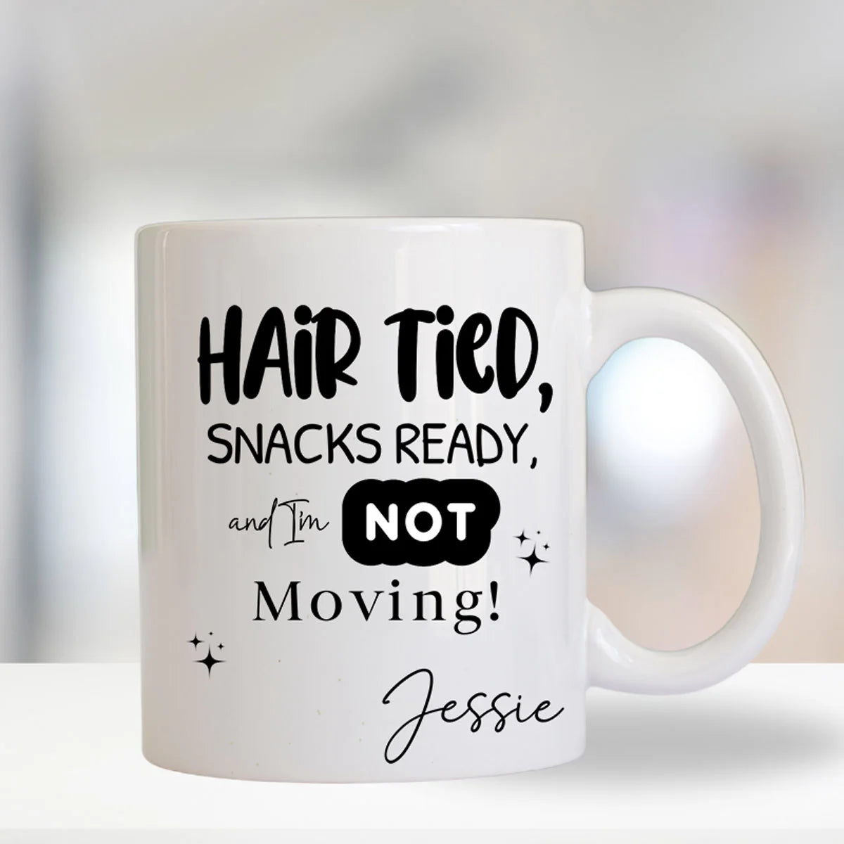White mug with black text 'Hair Tied, Snacks Ready, and I'm NOT Moving! Jessie' on a blurred background