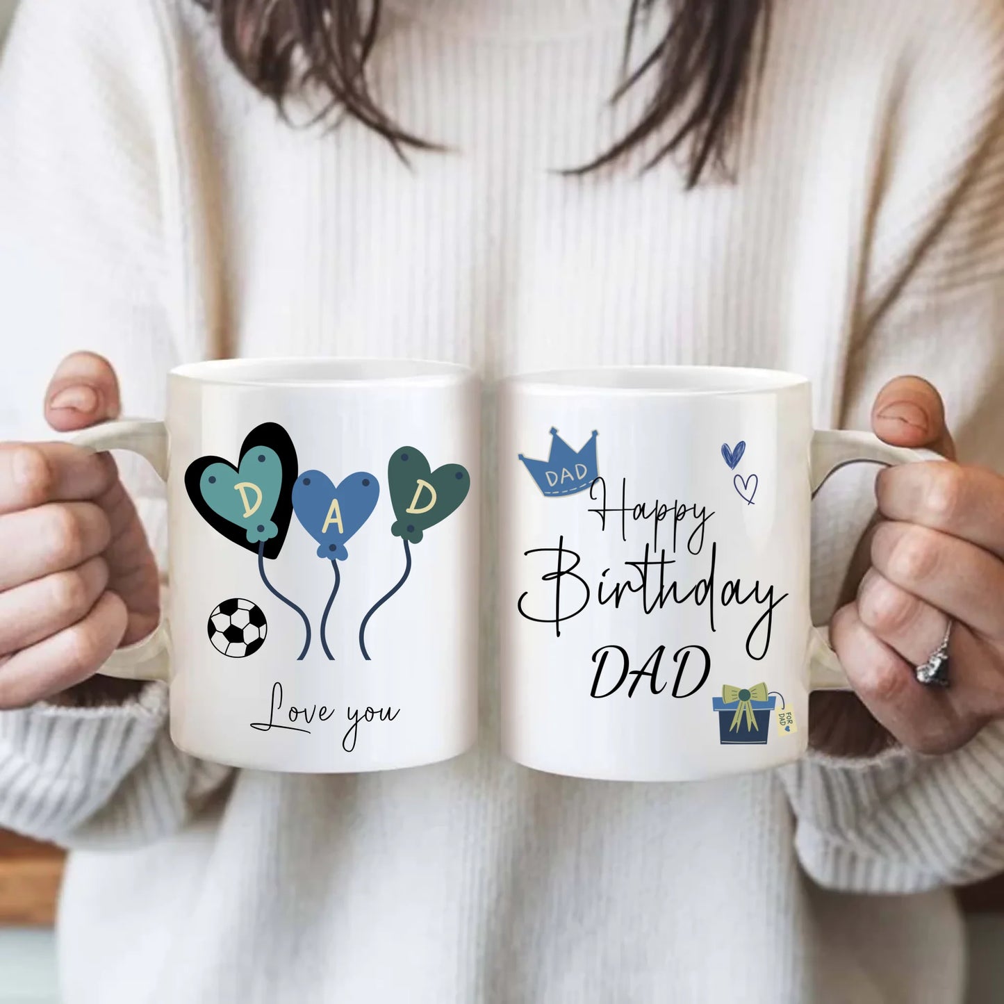 Two mugs with birthday-themed designs for a father, held by a person wearing a light sweater.