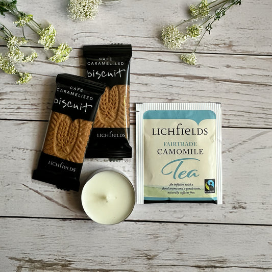 quirky-get-well-soon-letterbox-gift-uk. Two packages of Lichfields biscuit and tea on a wooden surface with a candle and flowers.