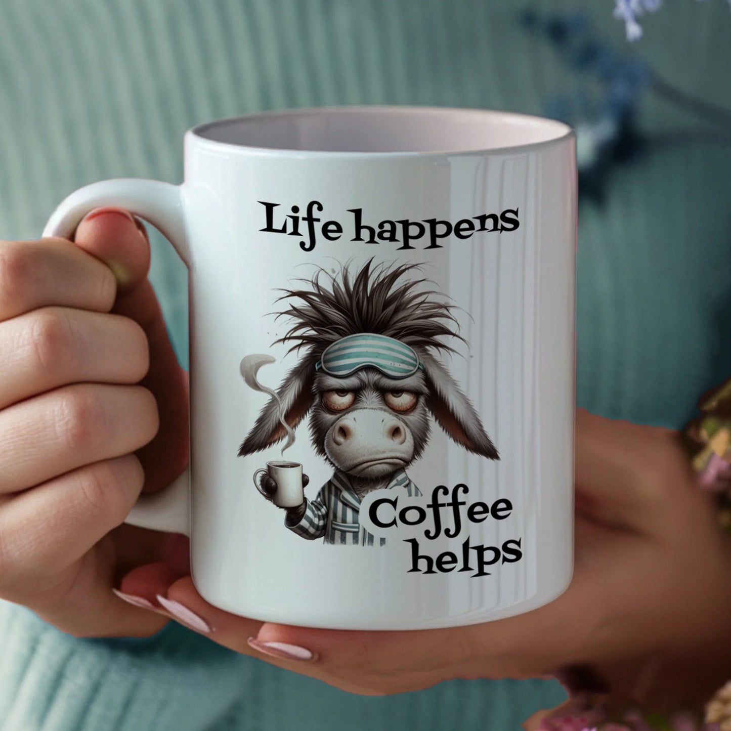 Person holding a mug with a cartoon donkey and text 'Life happens, Coffee helps' against a blurred background.