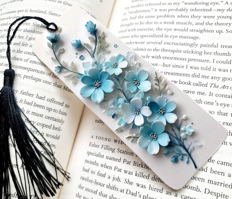 A pretty Bookmark made of mettal, printed with blue and white 3D flowers, giving a very pretty design, The bookmark has a white background, and is sitting on a page of wriiting.