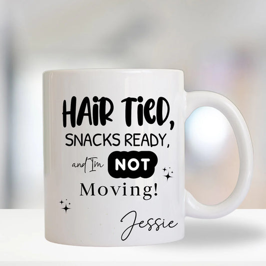 White mug with black text 'Hair Tied, Snacks Ready, and I'm NOT Moving! Jessie' on a blurred background