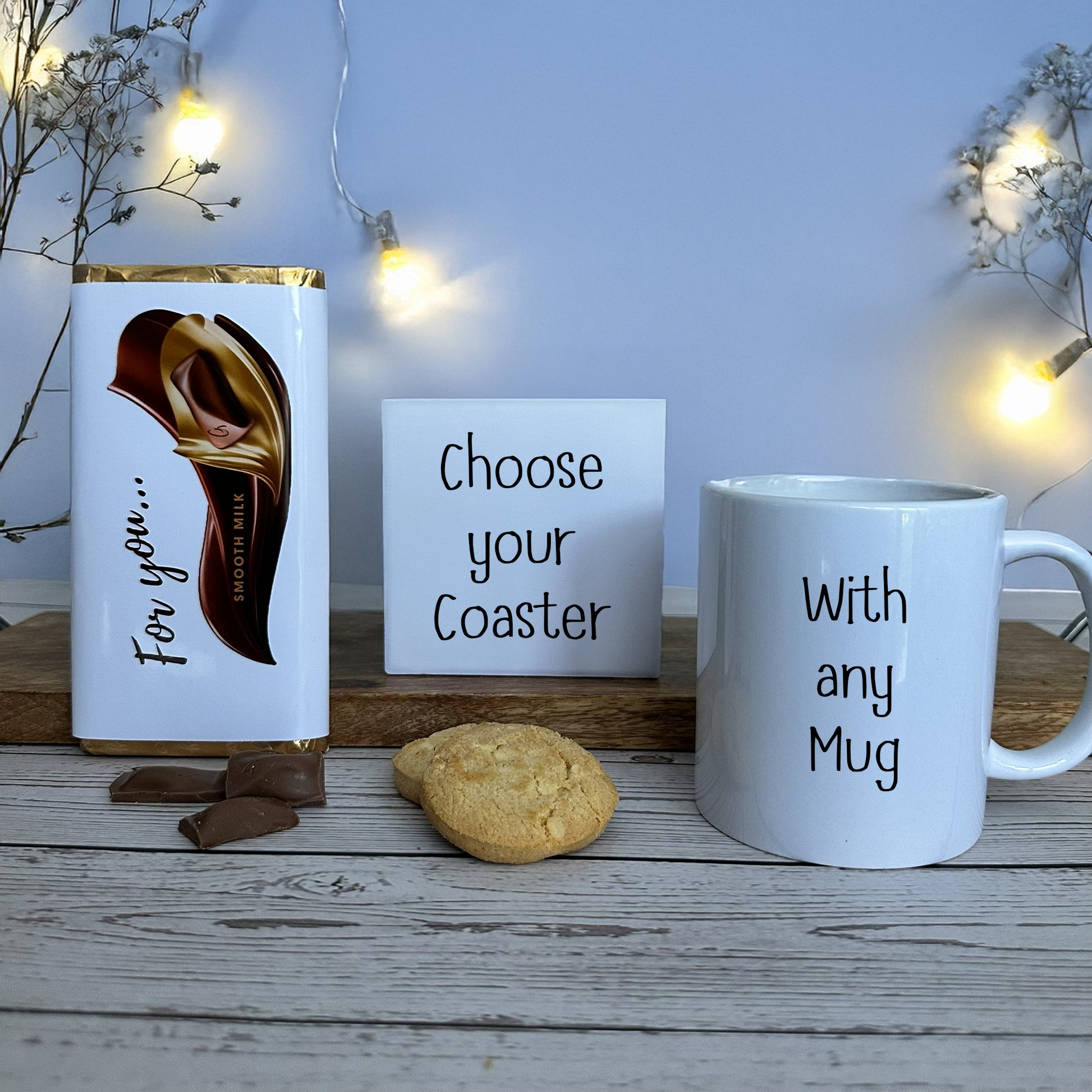 Create your own gift set - mug, chocolate bar, coaster, and cookies on a wooden surface with fairy lights.