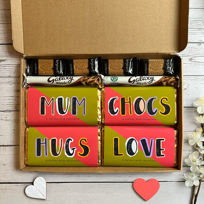 Box of Galaxy chocolate bars with 'Mum Chocs', 'Hugs', and 'Love' text on a wooden surface.