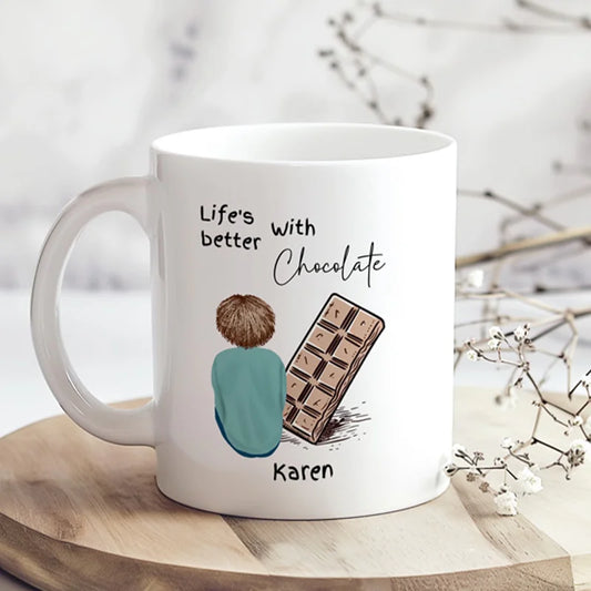 White mug with a design of a person and a chocolate bar, text 'Life's better with chocolate Karen', on a wooden surface with a blurred natural background.
