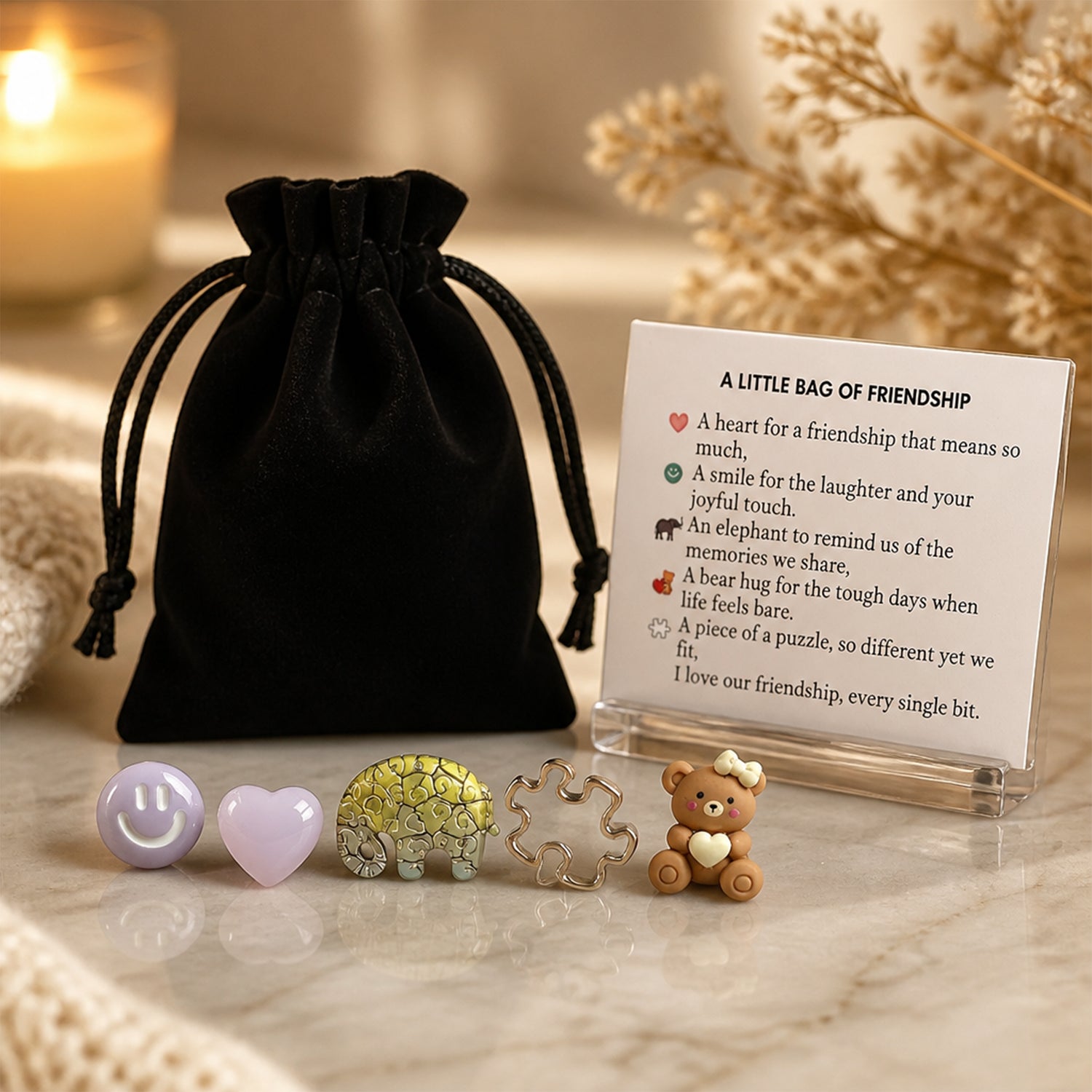 A friendship keepsake gift set displayed on a marble surface. The set includes a small black velvet drawstring pouch, a printed card titled 'A LITTLE BAG OF FRIENDSHIP and a beautiful verse about friendship -  a heart-shaped stone, a smiley face, a mosaic elephant charm, a silver puzzle piece, and a small brown teddy bear charm holding a white heart.