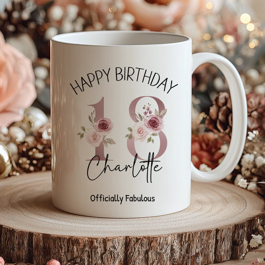 White mug with 'Happy Birthday Charlotte Officially Fabulous' text on a wooden surface with flowers in the background