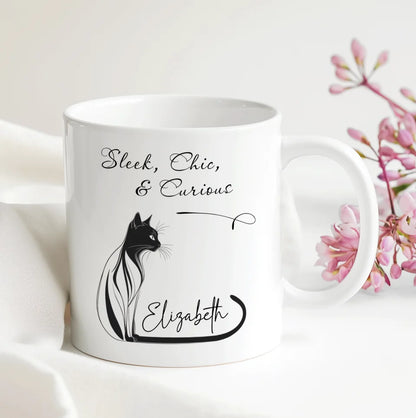 White mug with black cat illustration and text on a white background with pink flowers.