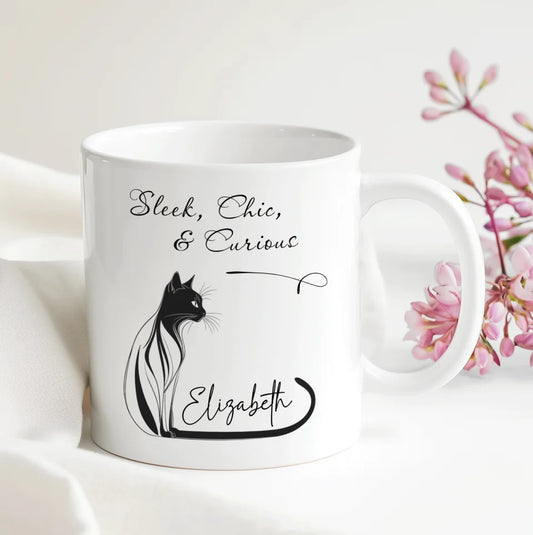 White mug with black cat illustration and text on a white background with pink flowers.