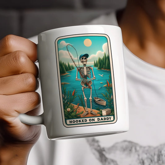 Mug with a skeleton fishing design and 'Hooked on Daddy' text, held by a person.